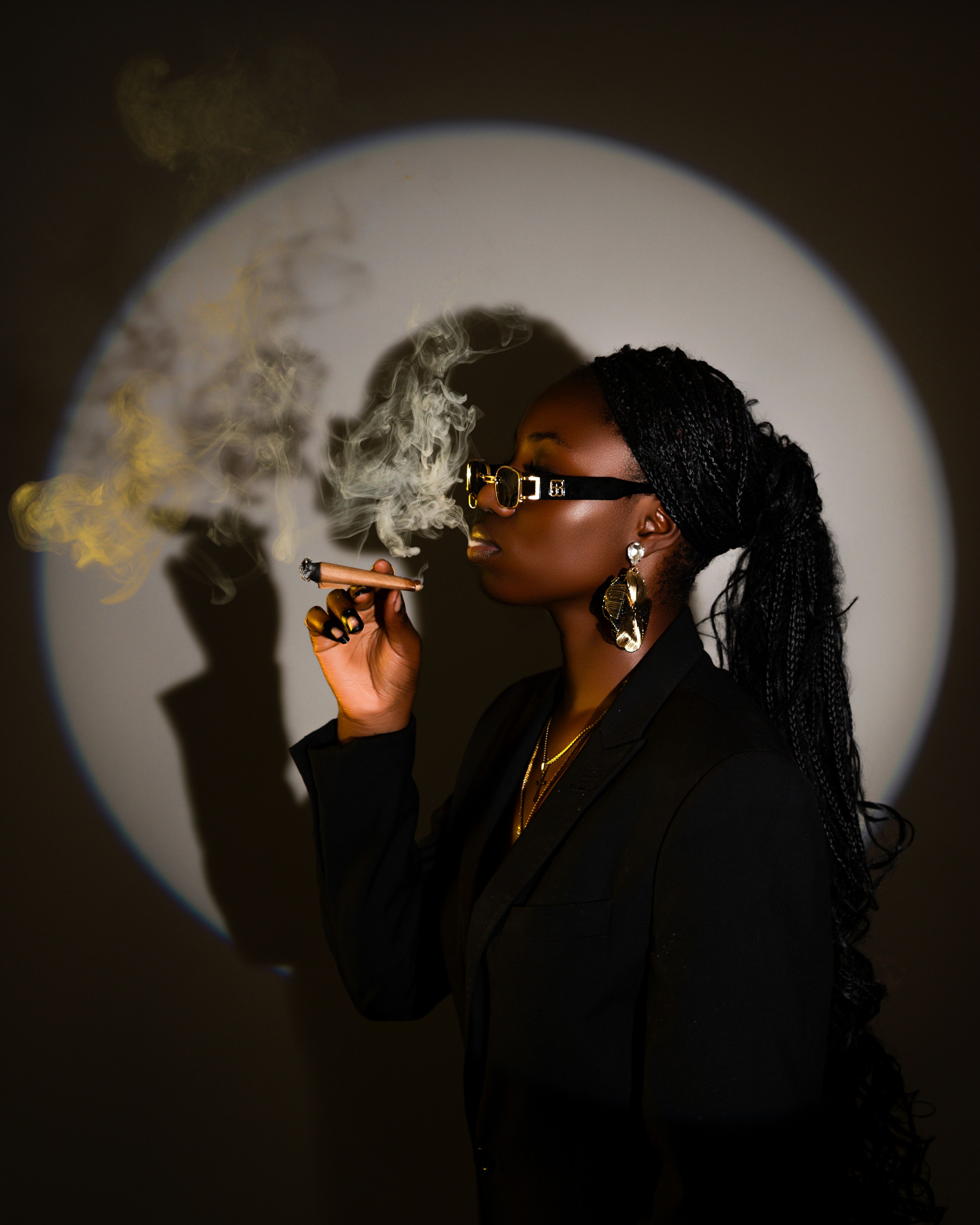 Chic woman in a dark suit and sunglasses elegantly smoking a cigar, surrounded by dramatic lighting and shadow.