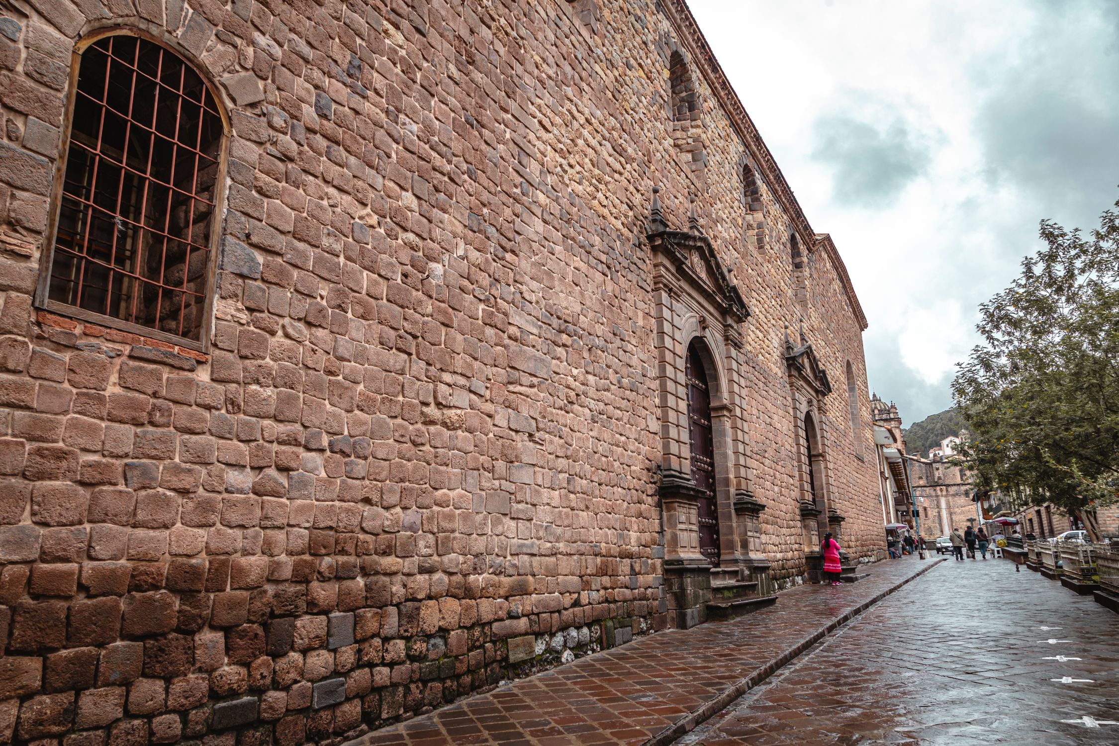 Brick wall and street scene in historic Cusco, Peru with people and a cloudy sky.