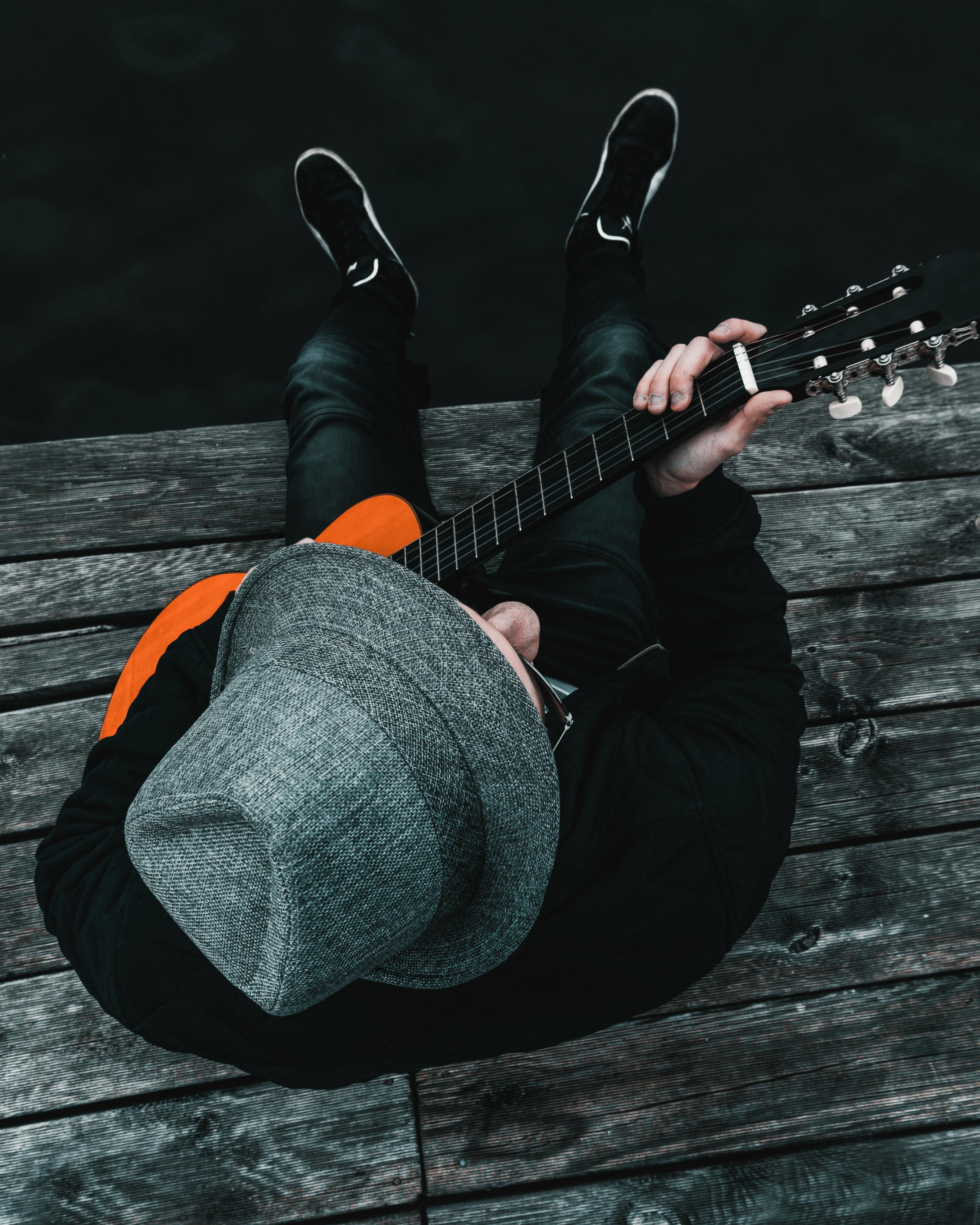 一位戴着软毡帽、背着吉他的音乐家正坐在木码头上享受休闲时光