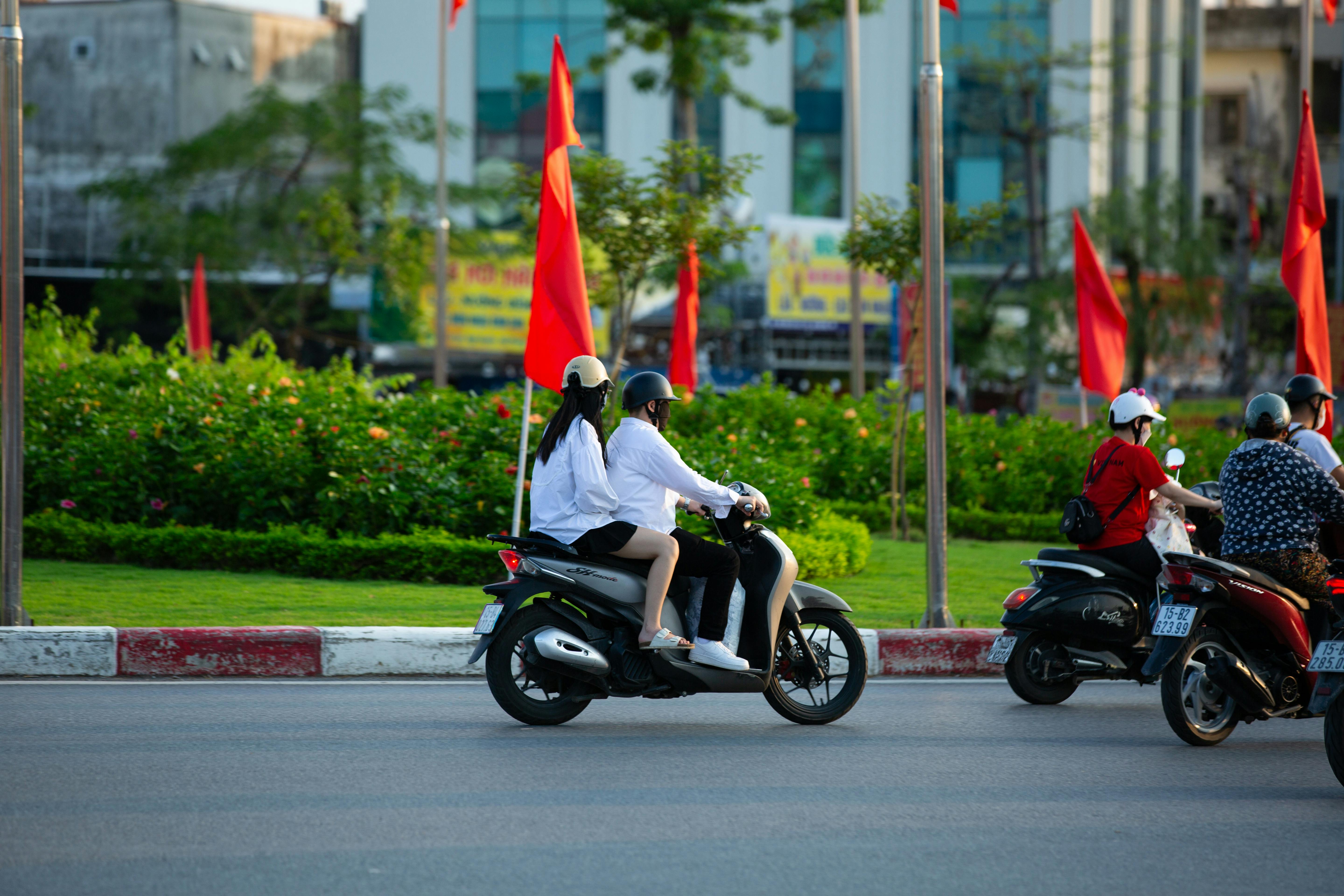 A group of people riding scooters on an urban road lined with red flags, indicating a vibrant city scene.