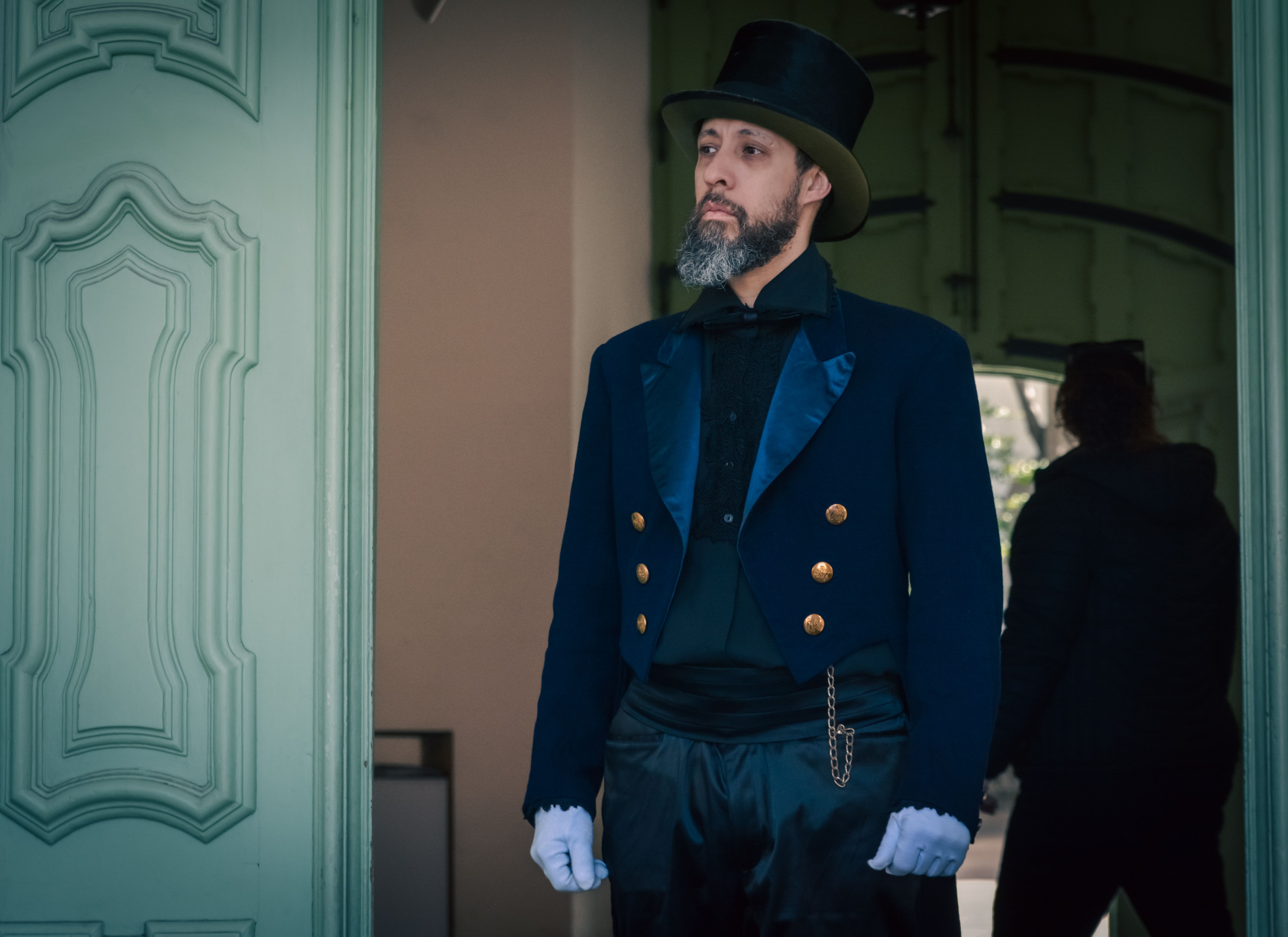 A man in elegant vintage clothing stands by an intricately designed door indoors.
