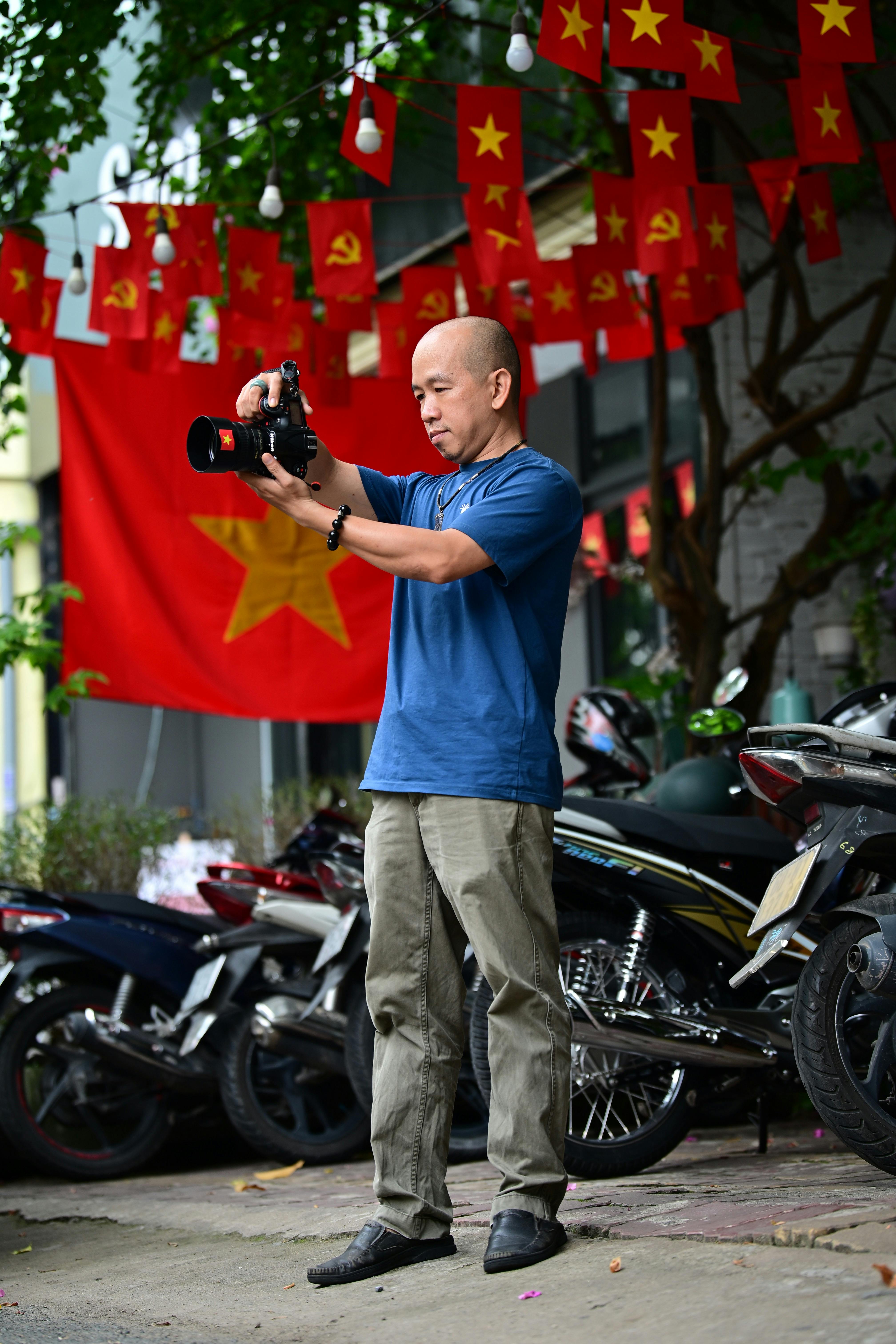 Man taking photo with camera outdoors, flags in background, motorcycles nearby.