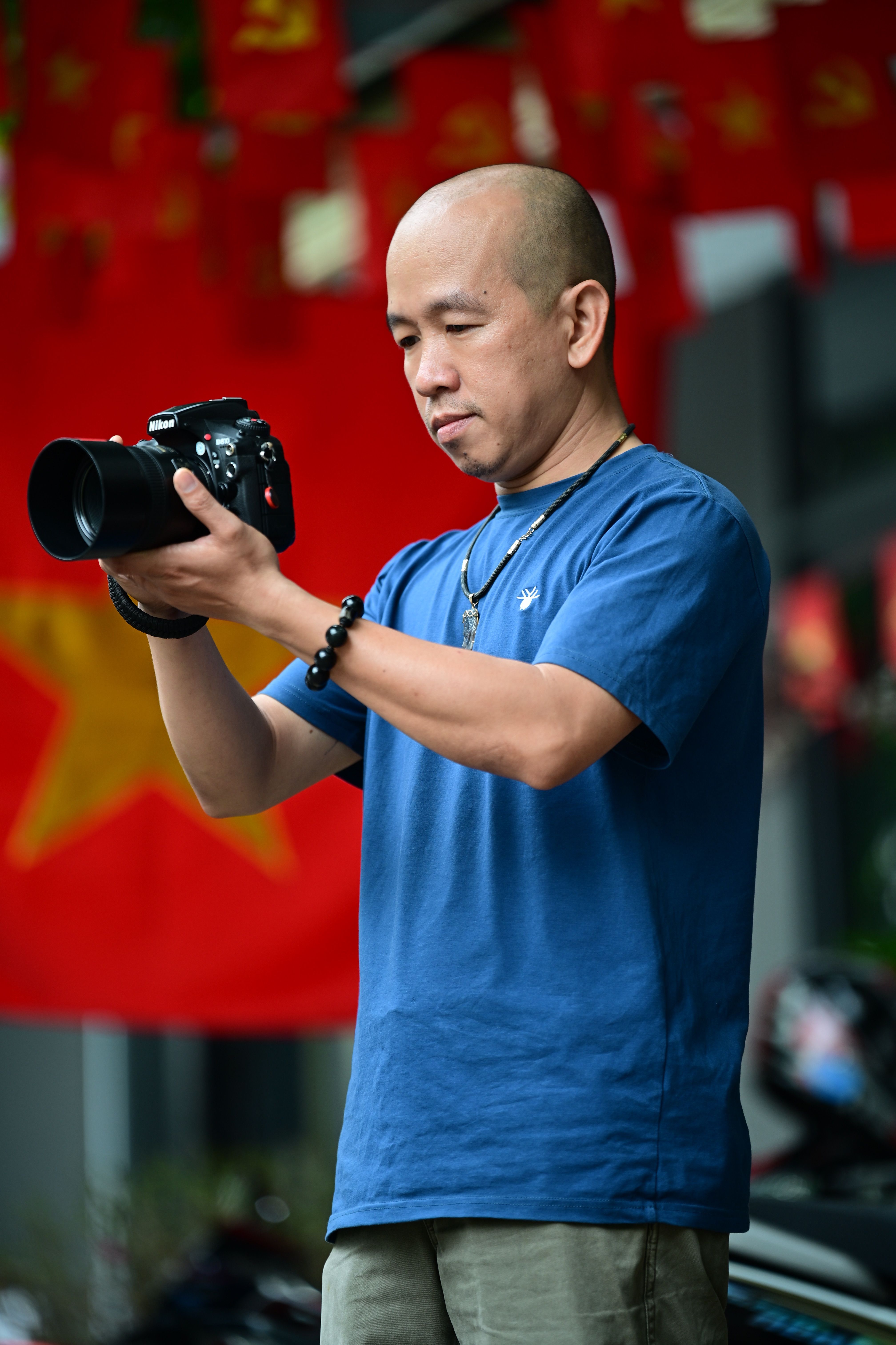 A photographer in a blue shirt adjusts a camera surrounded by vibrant red flags, capturing a moment outdoors.