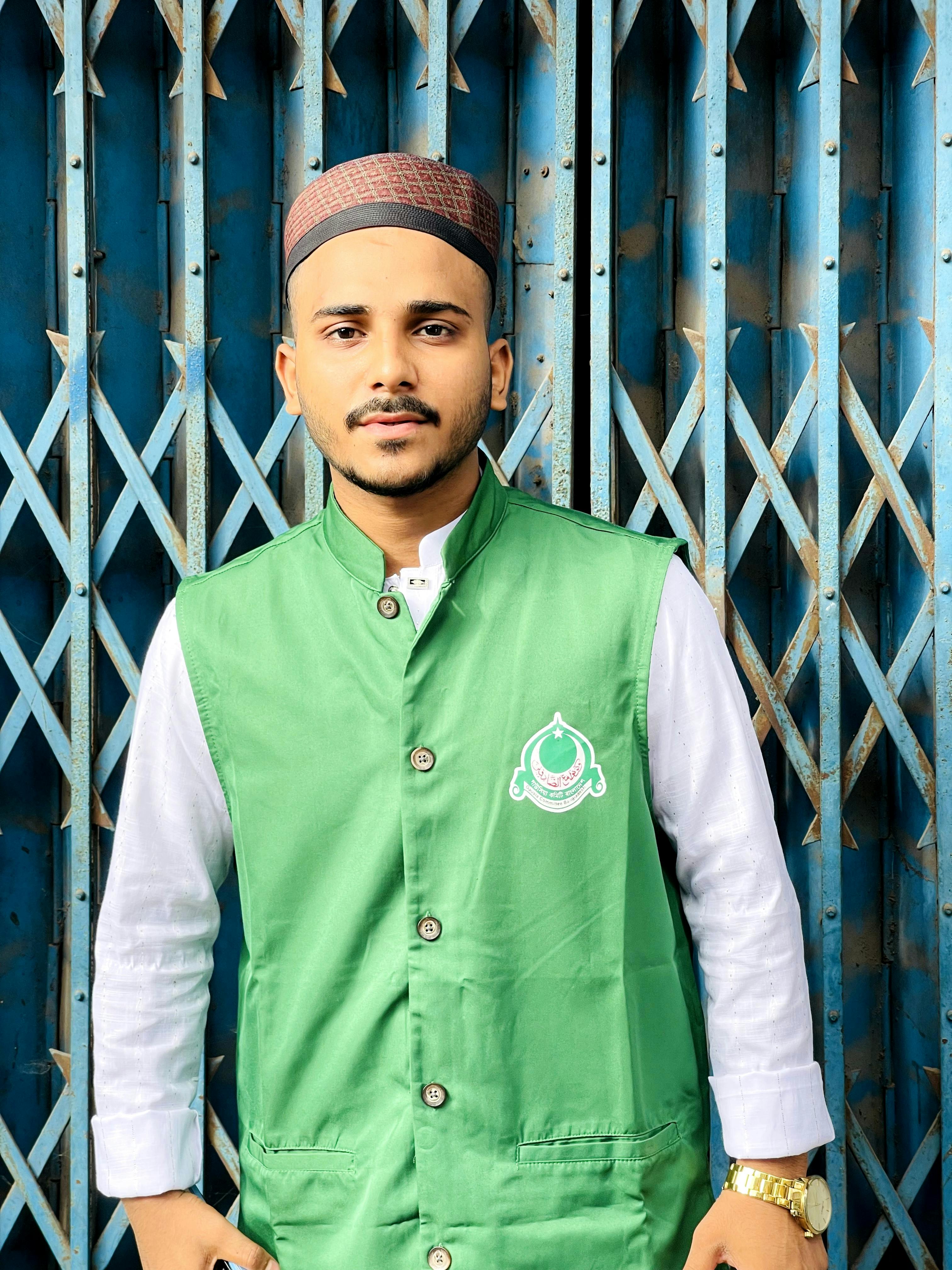 Young adult male wearing traditional attire stands confidently in front of metal gate.