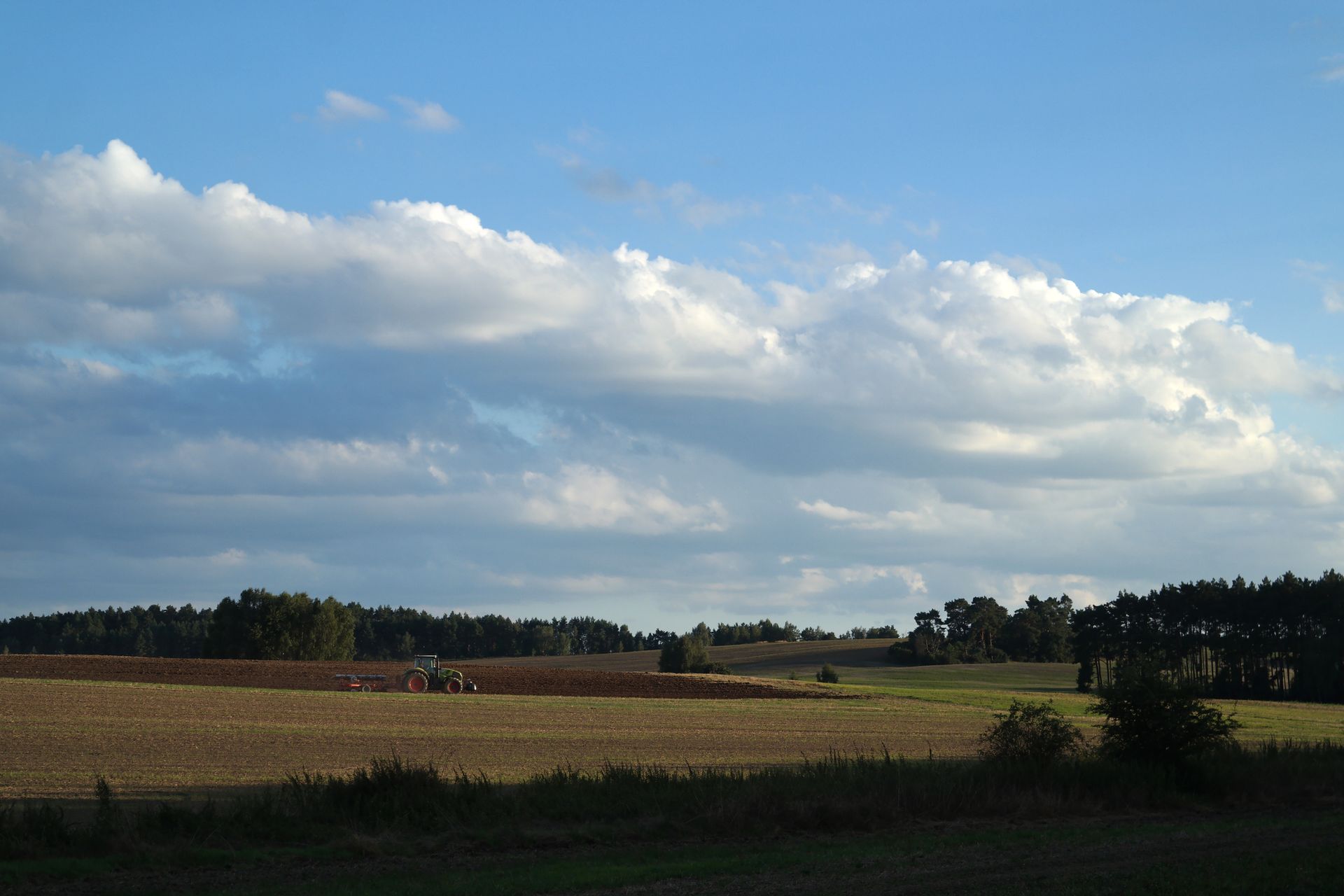 A peaceful rural scene featuring a tractor working in a vast agricultural field under a blue sky with clouds.