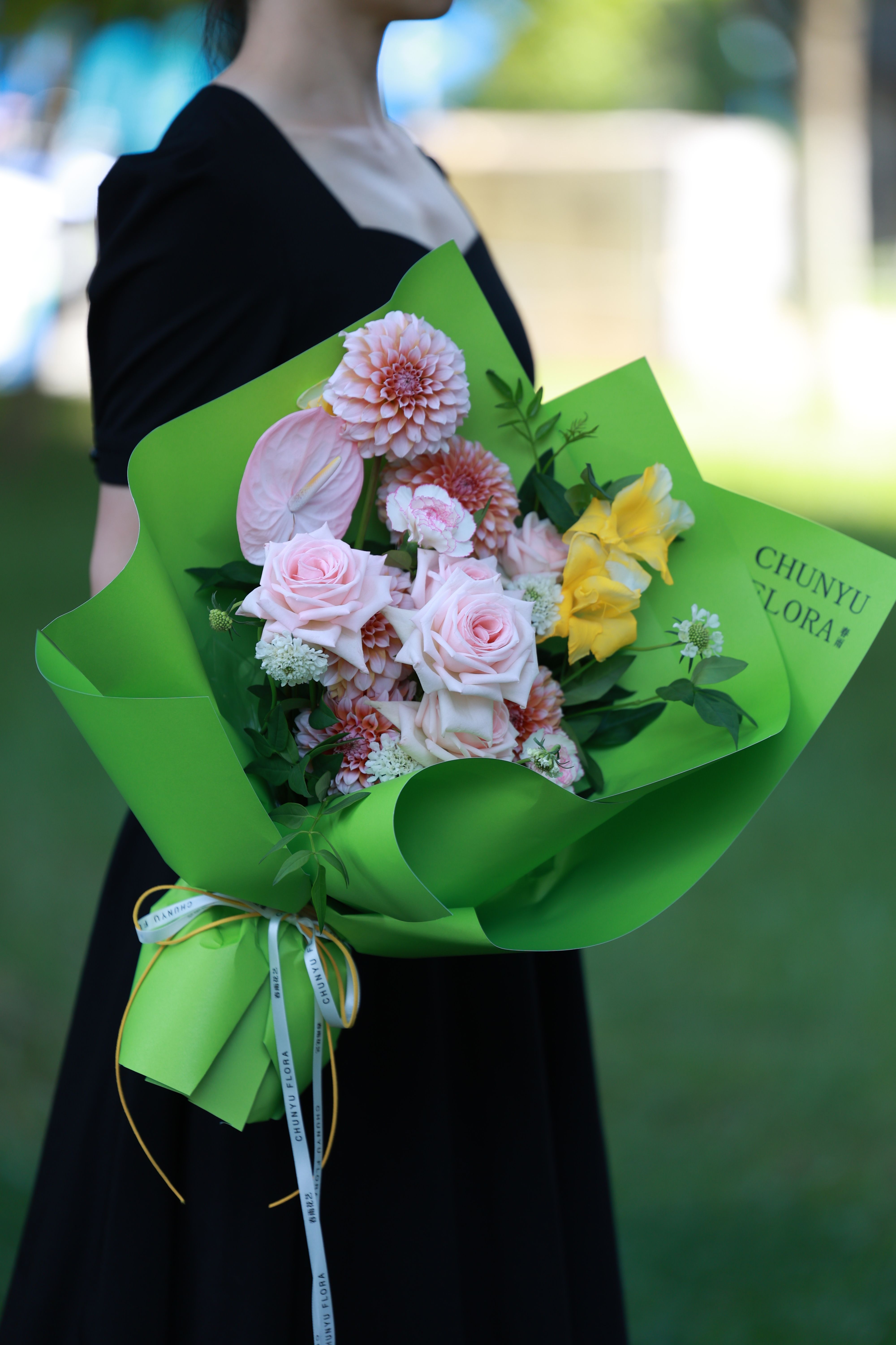 A beautiful bouquet of roses and dahlias wrapped in green paper, held by a person in a black dress.