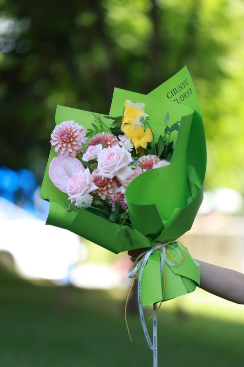Free A beautiful bouquet with pink and yellow flowers wrapped in green paper, held outdoors. Stock Photo