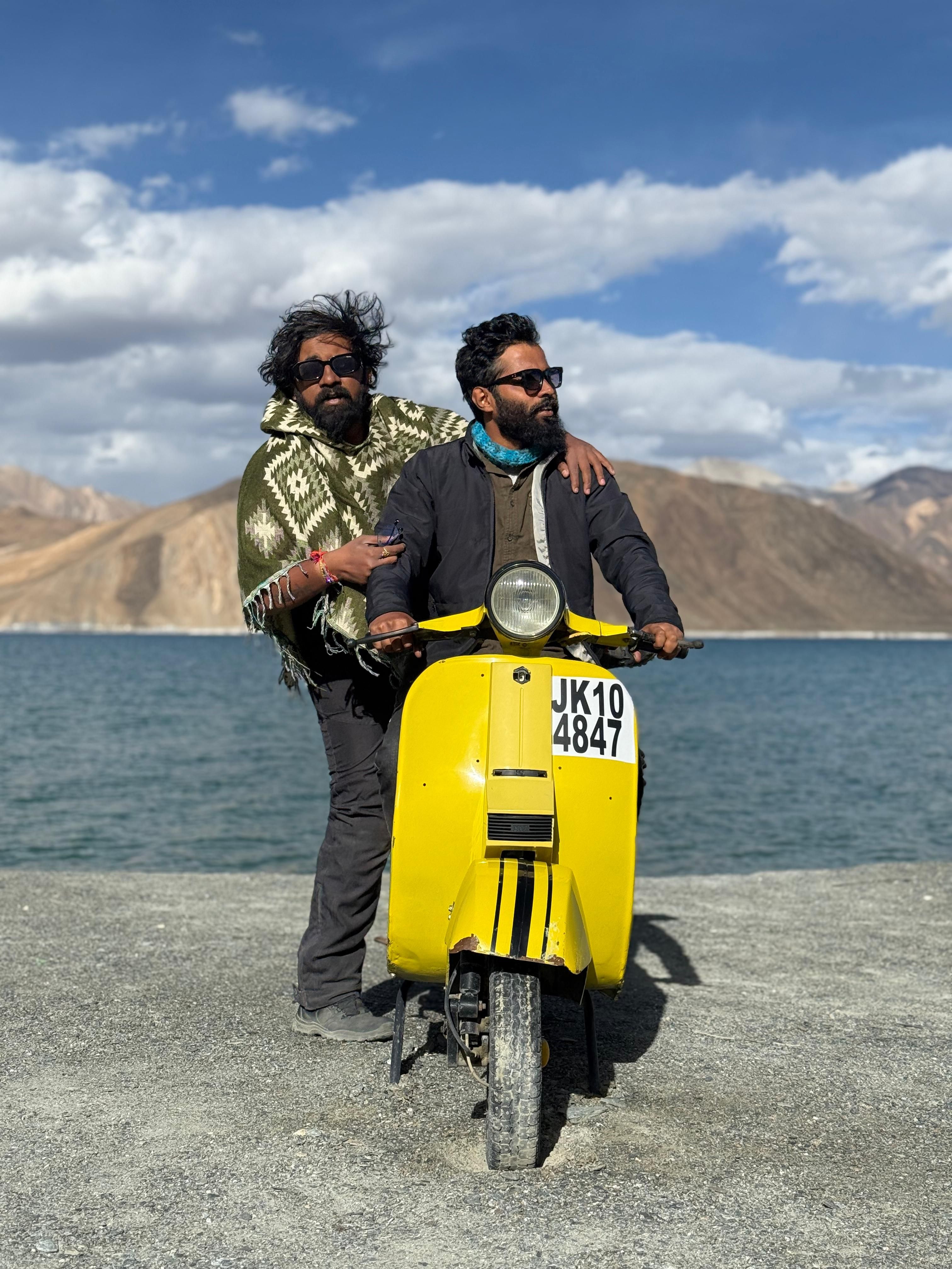 Two adventurers pose with a yellow scooter beside a scenic lake in Leh.
