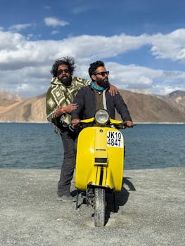 Two adventurers pose with a yellow scooter beside a scenic lake in Leh.