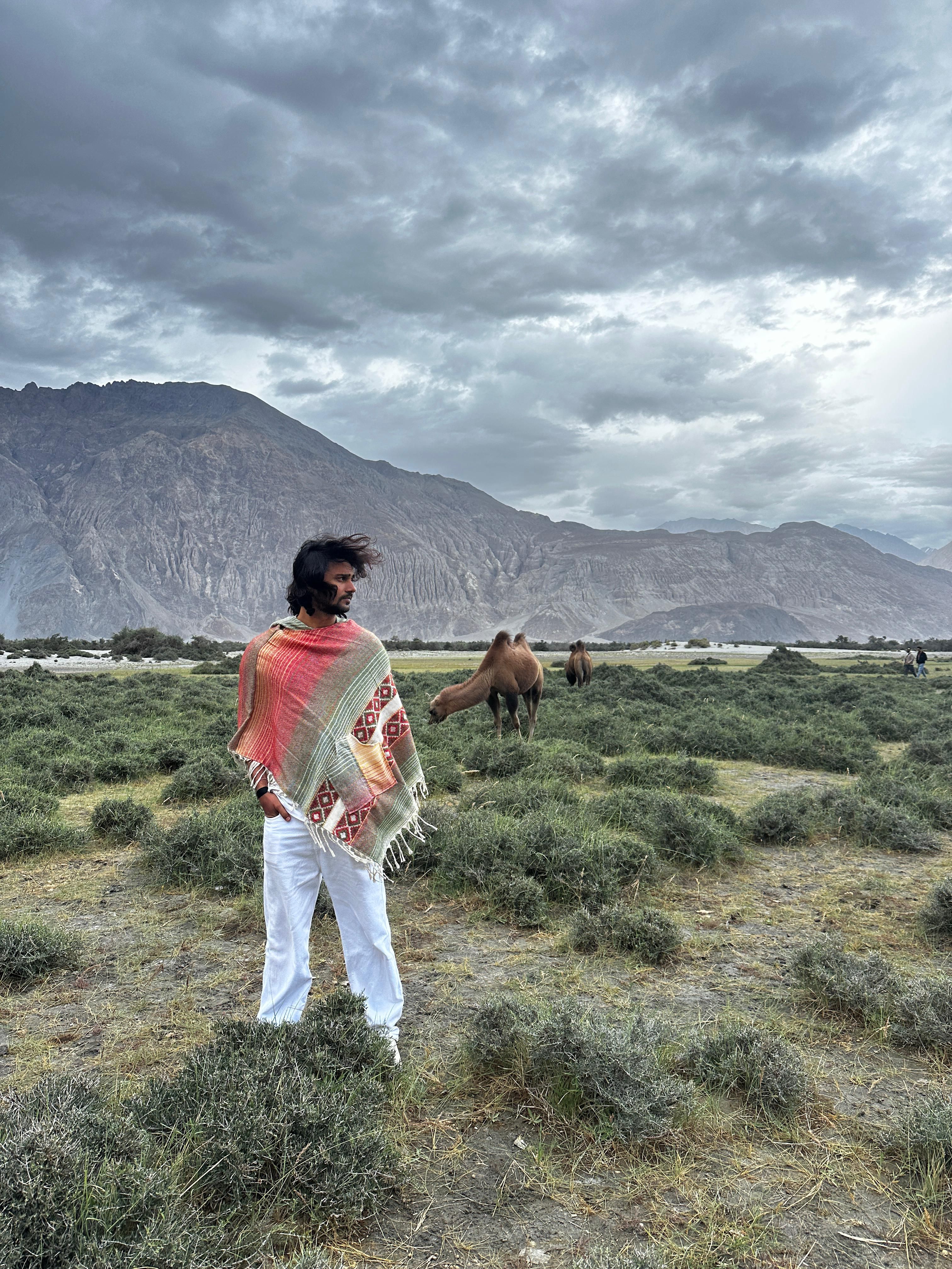 A man wrapped in a colorful shawl stands in a grassy landscape with camels, under dramatic skies.