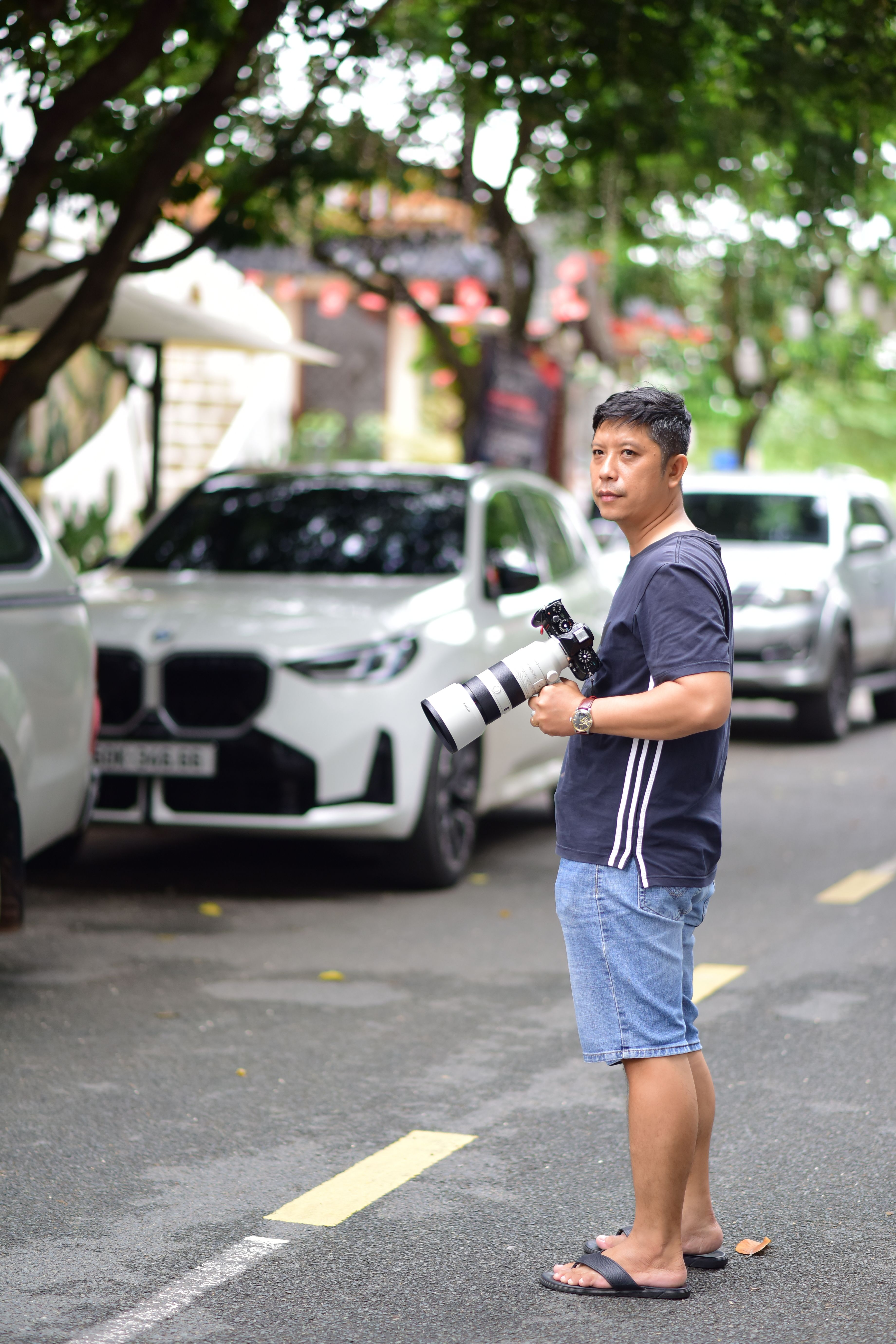 Photographer standing on a street holding a camera with a telephoto lens, showcasing casual urban photography style.