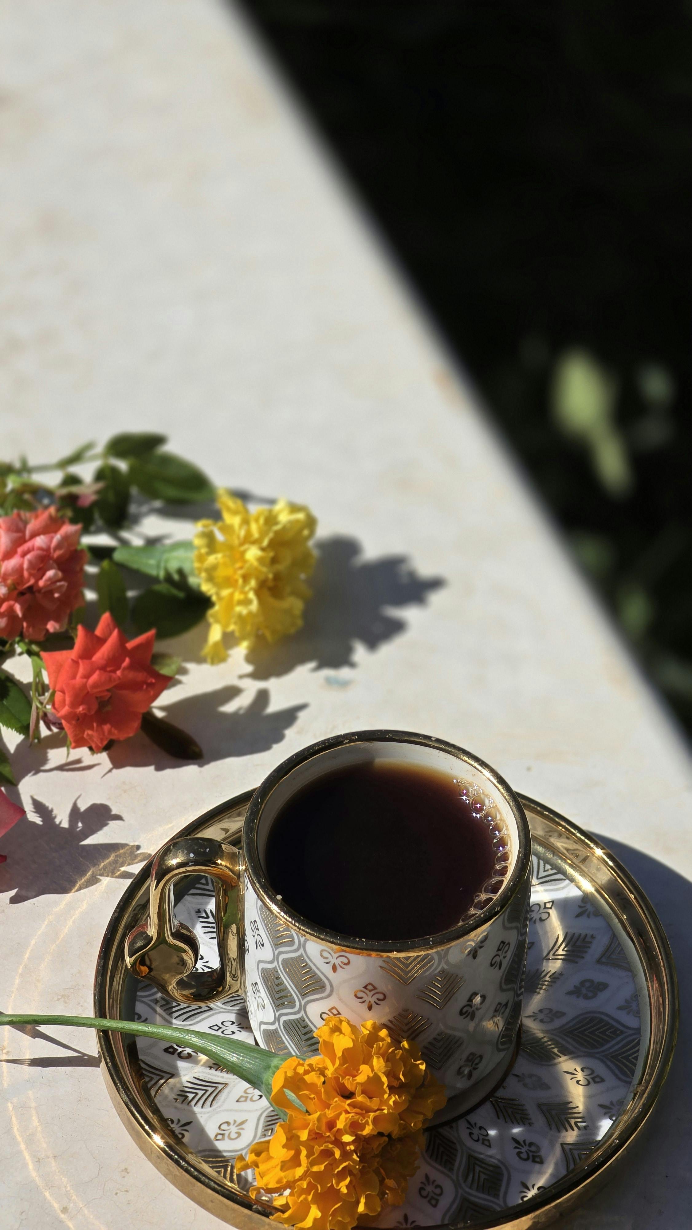 A elegantly detailed cup of black tea on a saucer with colorful flowers in sunlight.