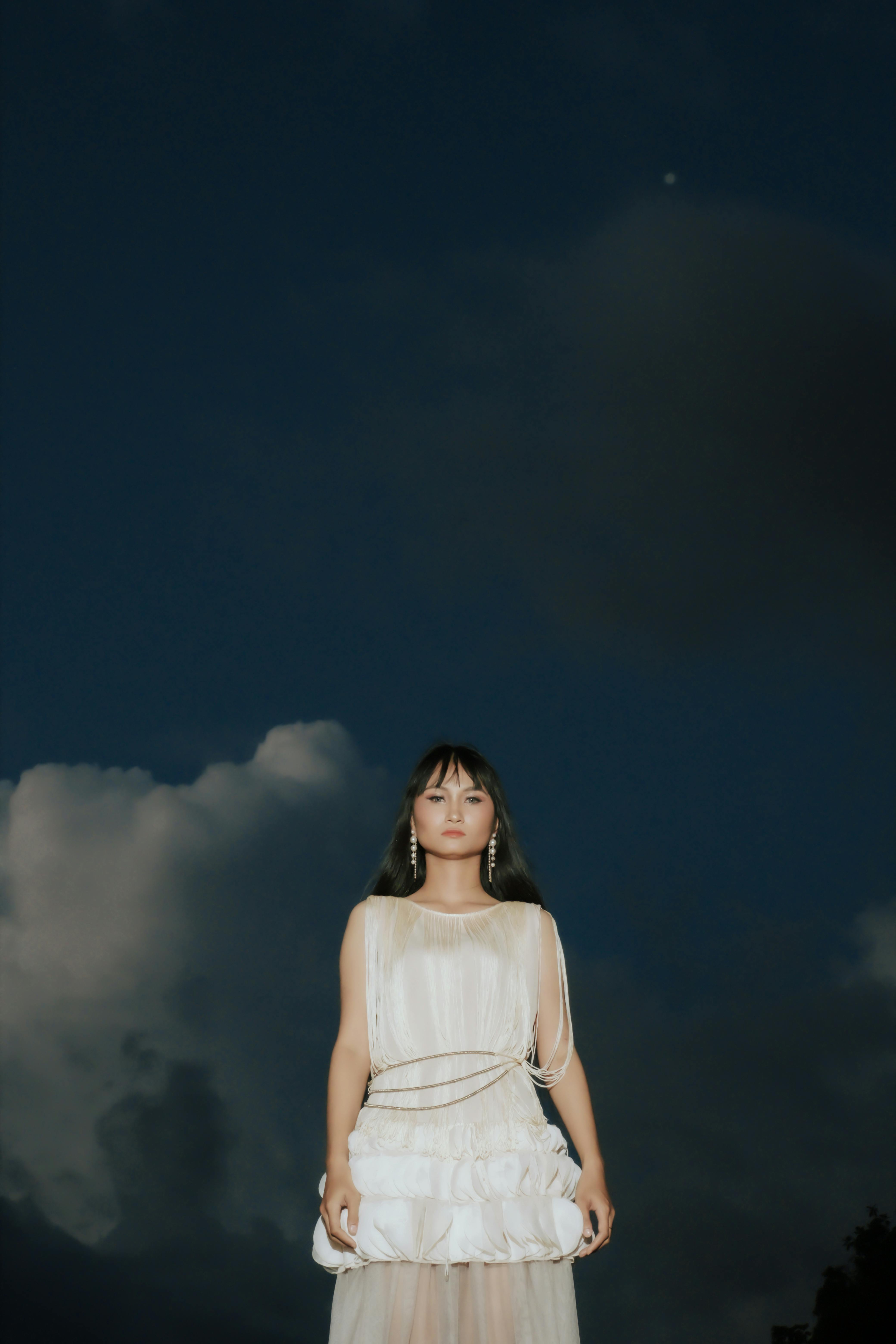 A serene portrait of a woman in a white dress against a dramatic dark sky, creating a moody atmosphere.