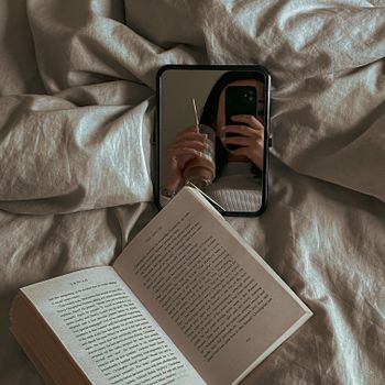 A cozy scene featuring a mirror selfie with an iced coffee and an open book on a bed.