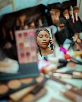 A woman applies makeup, reflected in a mirror surrounded by makeup brushes and cosmetics.