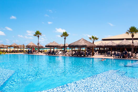 A beautiful tropical resort featuring a pool, thatched huts, and palm trees under a clear blue sky.