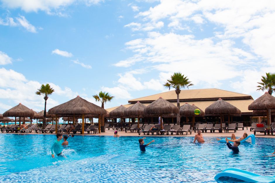 Famílias relaxando em uma piscina de resort de luxo com palmeiras e guarda-sóis, destacando atividades familiares.