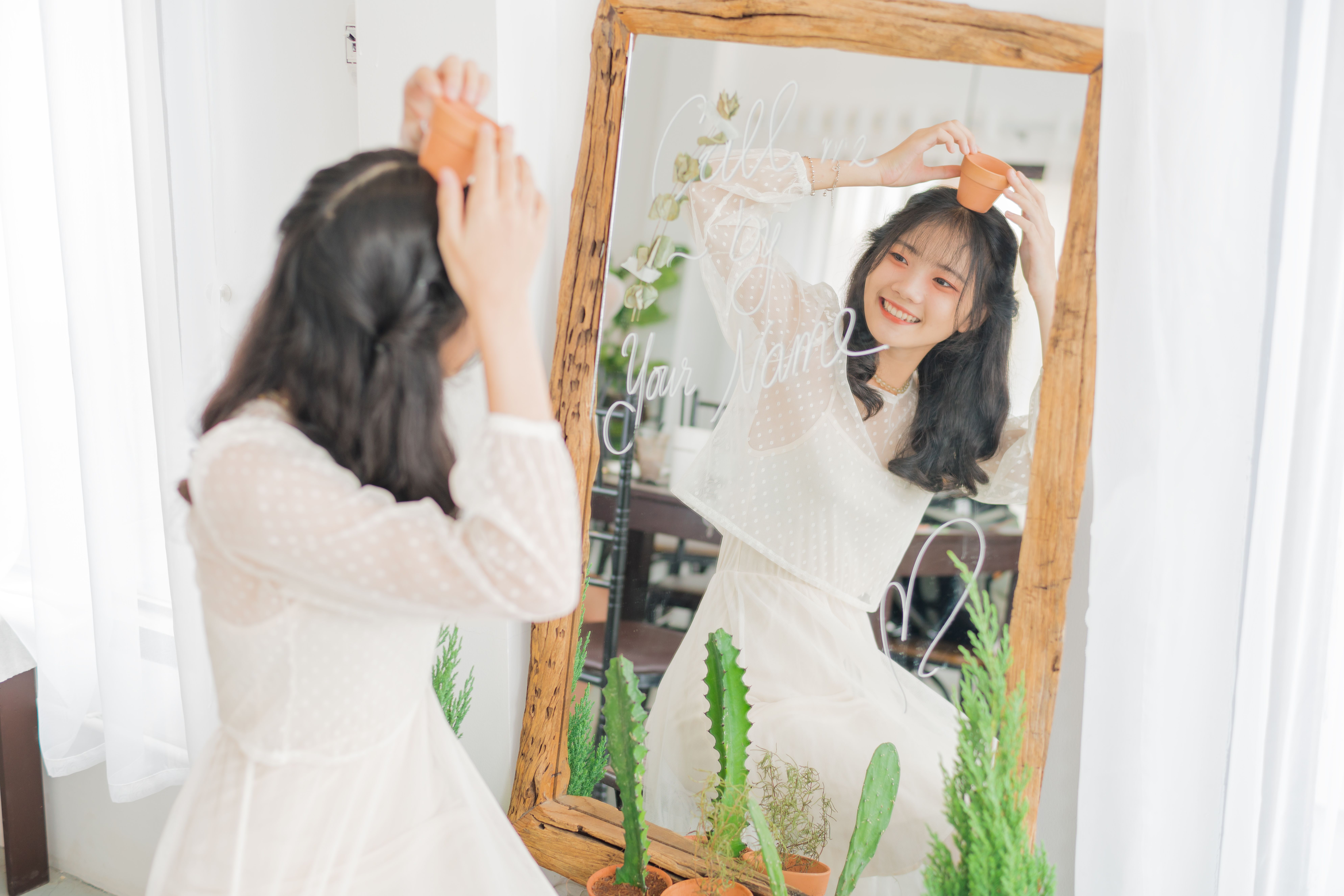 Smiling woman holding a small pot in front of mirror with plants, embracing nature indoors.
