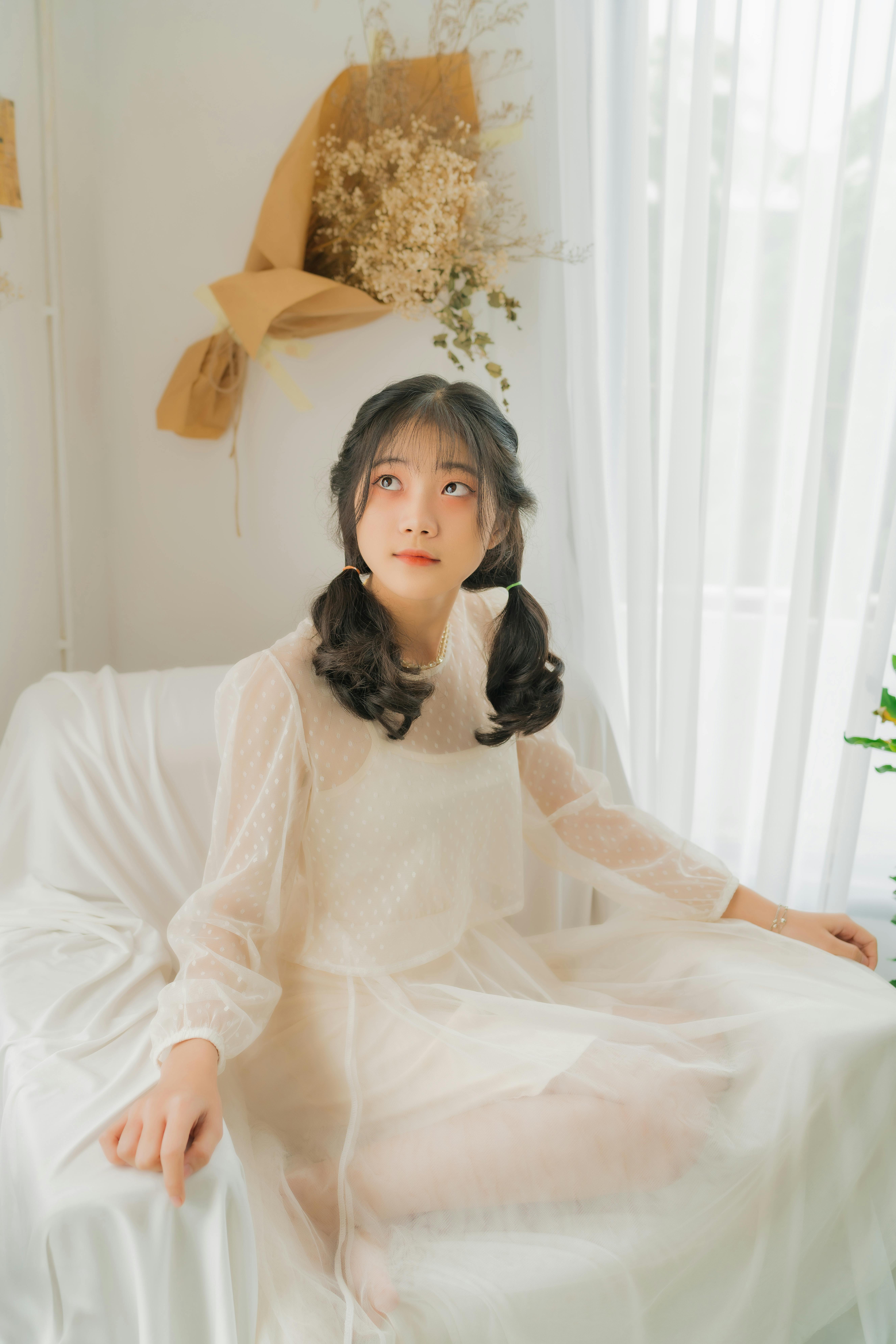 A young woman in a delicate white dress sits gracefully on a chair by the window.