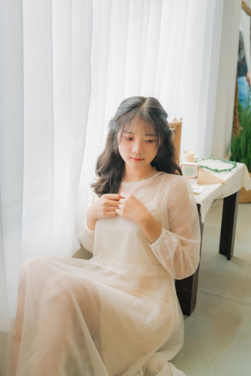 Free Elegant woman in white dress sitting by window in soft natural lighting. Stock Photo