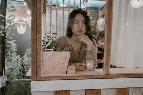 Free A young woman sitting thoughtfully at a cafe, gazing through the window with plants around her. Stock Photo