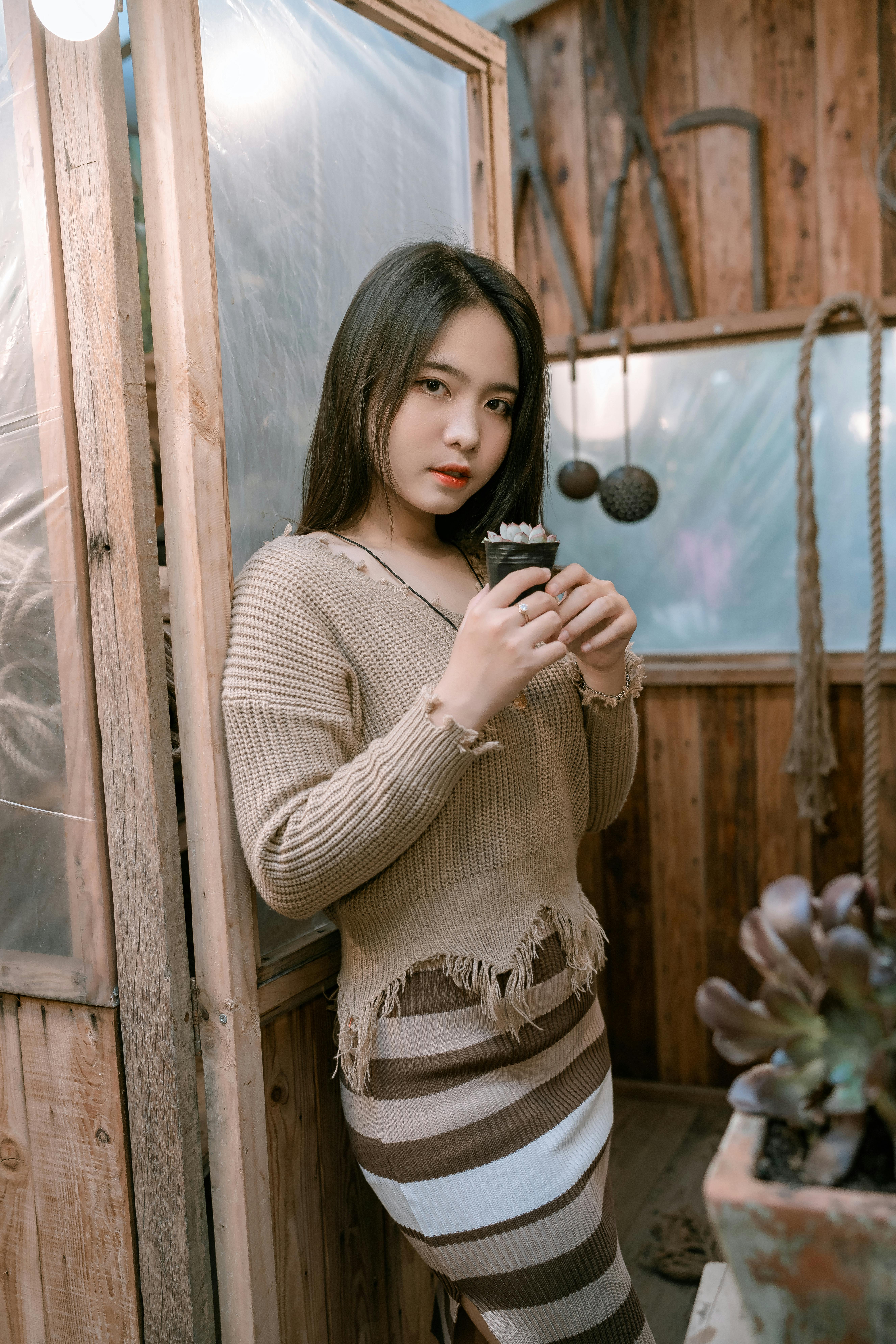 Young woman indoors holding a small plant, showcasing cozy fashion and home decor.