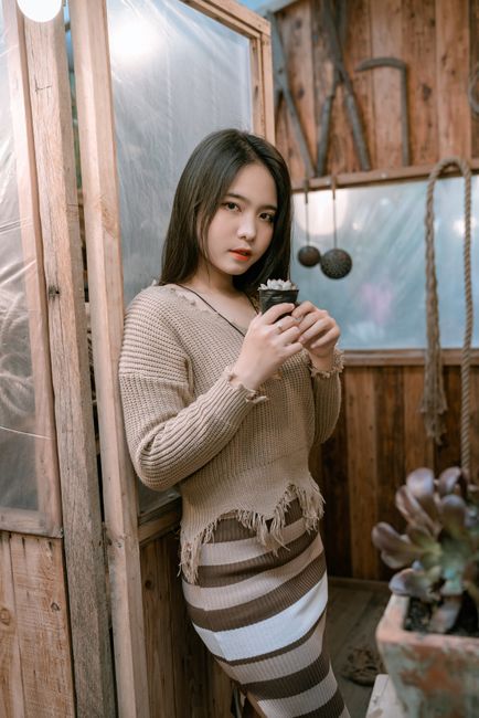 Young woman indoors holding a small plant, showcasing cozy fashion and home decor.