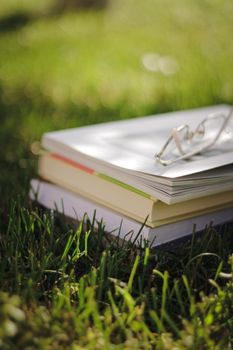 A stack of books with reading glasses rests on green grass under sunlight, evoking summer learning.