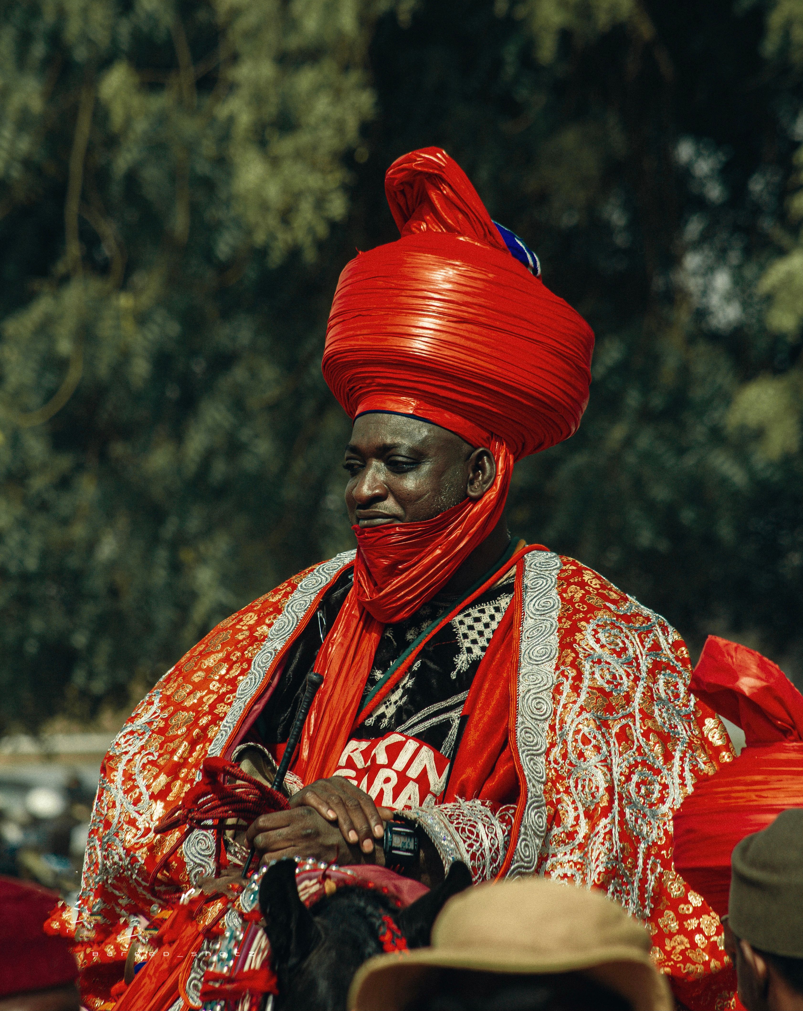 A man in regal Nigerian outfit during a cultural festival outdoors, rich in red and intricate patterns.