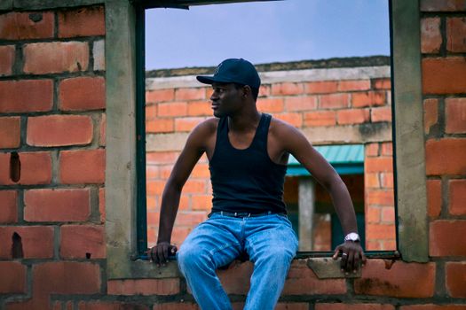 A young man in casual attire sits in a window frame of a brick building.