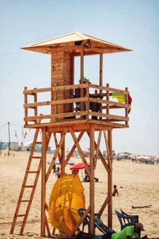 A wooden lifeguard tower stands on a sandy beach with people enjoying a sunny day.