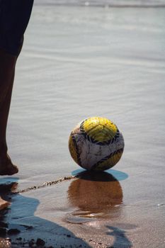 A colorful soccer ball on a beach with a person's leg and reflection in the wet sand.