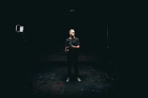 Free A cinematographer standing on a dimly lit film set, holding camera equipment. Stock Photo