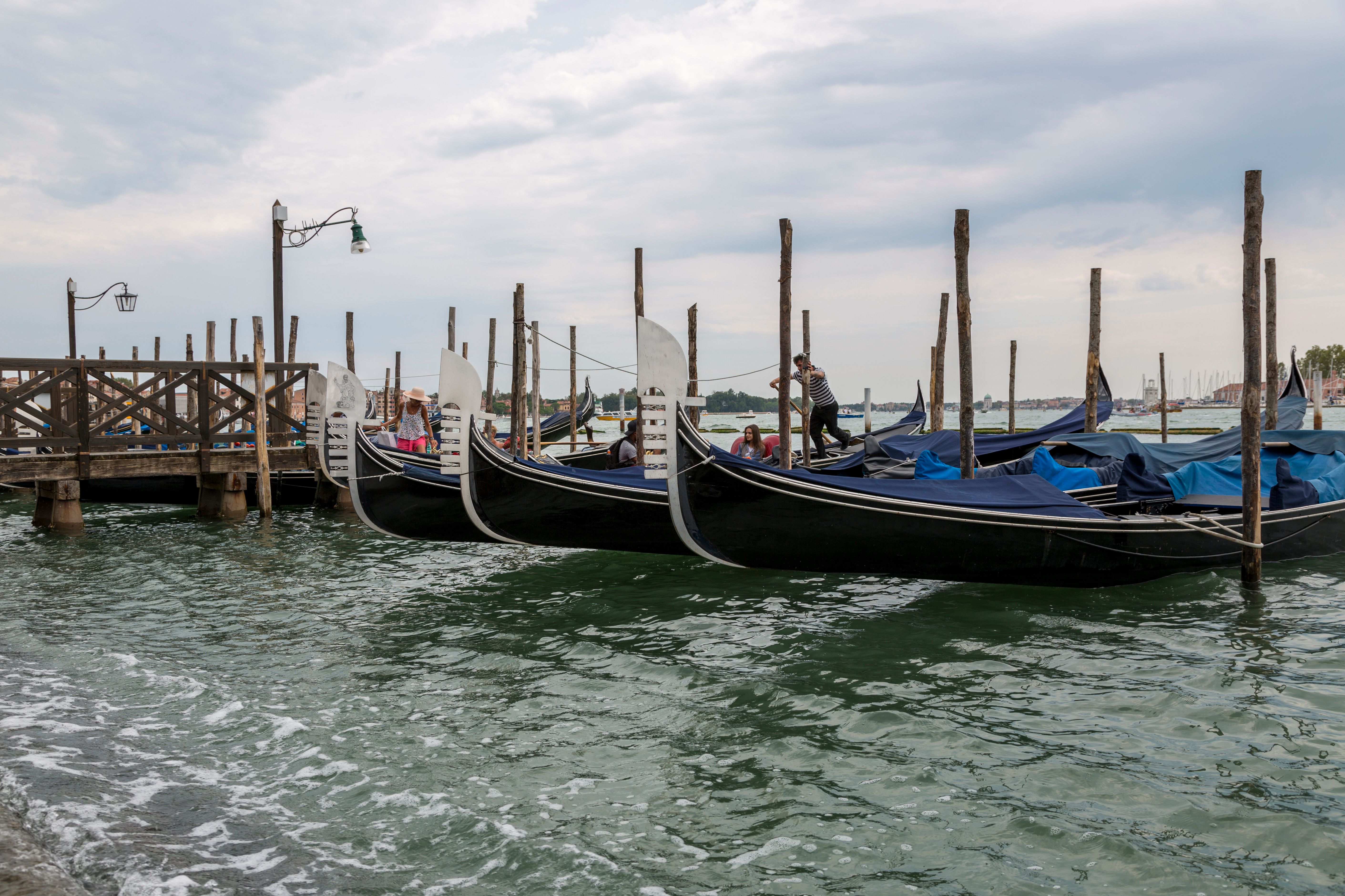 Gondolas docked at a pier in Venice, Italy with a cloudy sky above. Calm waters ripple against the boats.