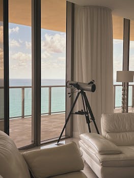 Modern living room with telescope facing a stunning beach view through large windows.