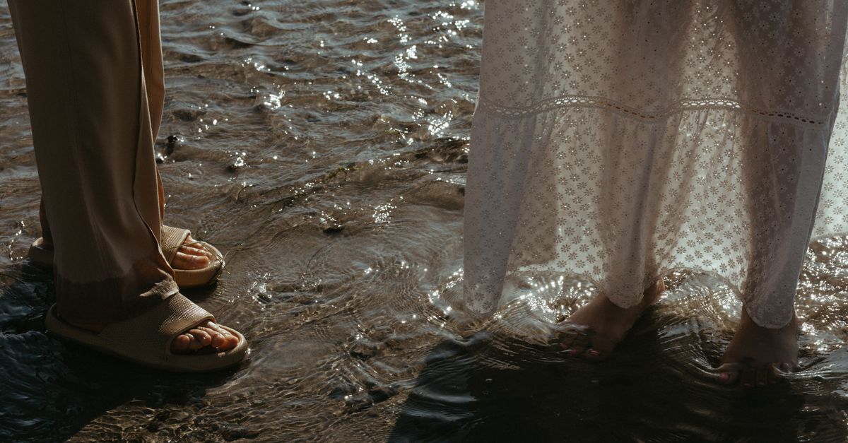 Feet dressed in sandals and a white dress standing at the water's edge on a sunny beach.