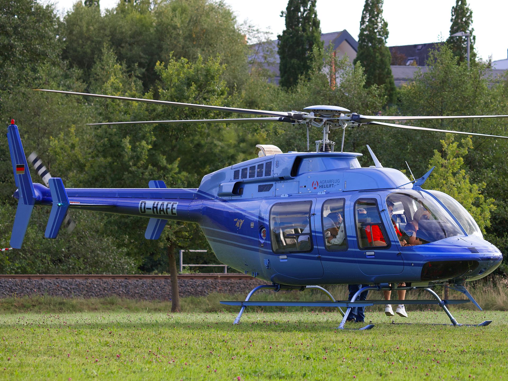 Blue helicopter parked on grassy field with pilot and passenger visible inside.