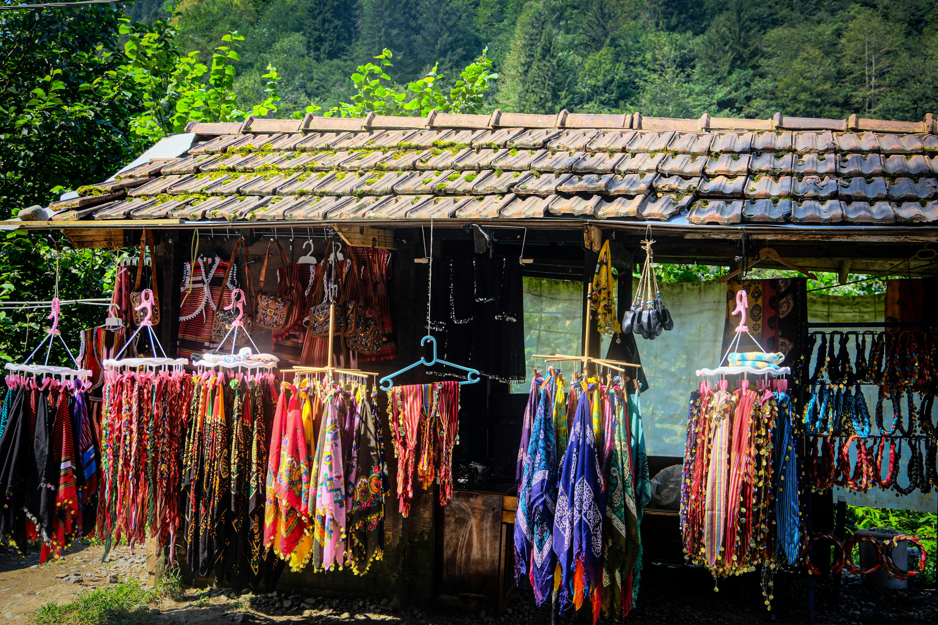 A vibrant outdoor market stall displaying colorful textiles and accessories against a lush forest backdrop.