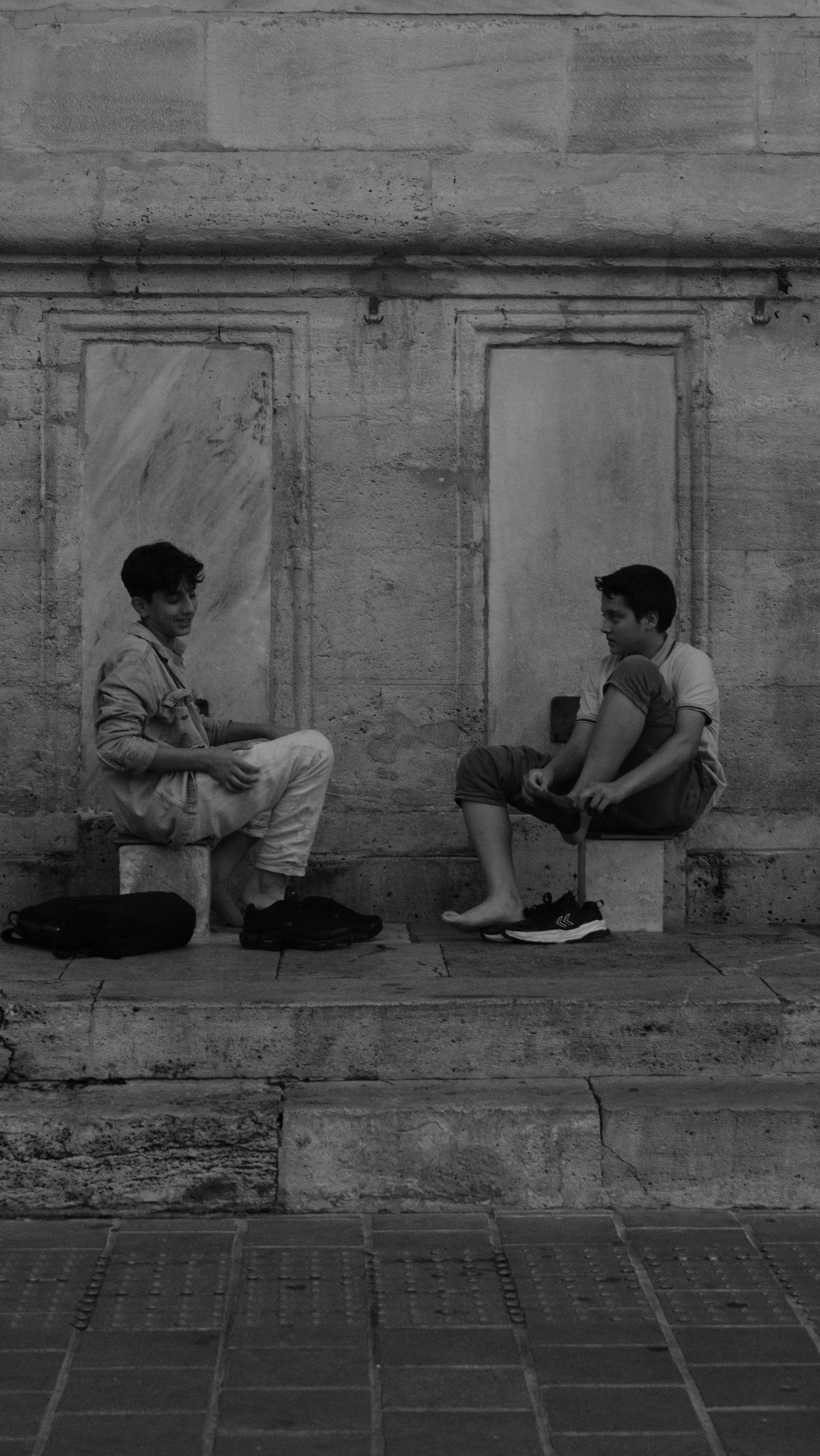 Gratis Due ragazzi sono seduti casualmente sui gradini, intenti a conversare, con uno sfondo di muro di pietra. Foto a disposizione