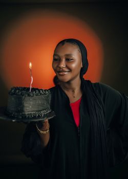 Smiling woman holding a black birthday cake with a lit candle, exuding joy and celebration.