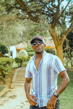 Man in sunglasses and striped shirt outdoors with greenery background.