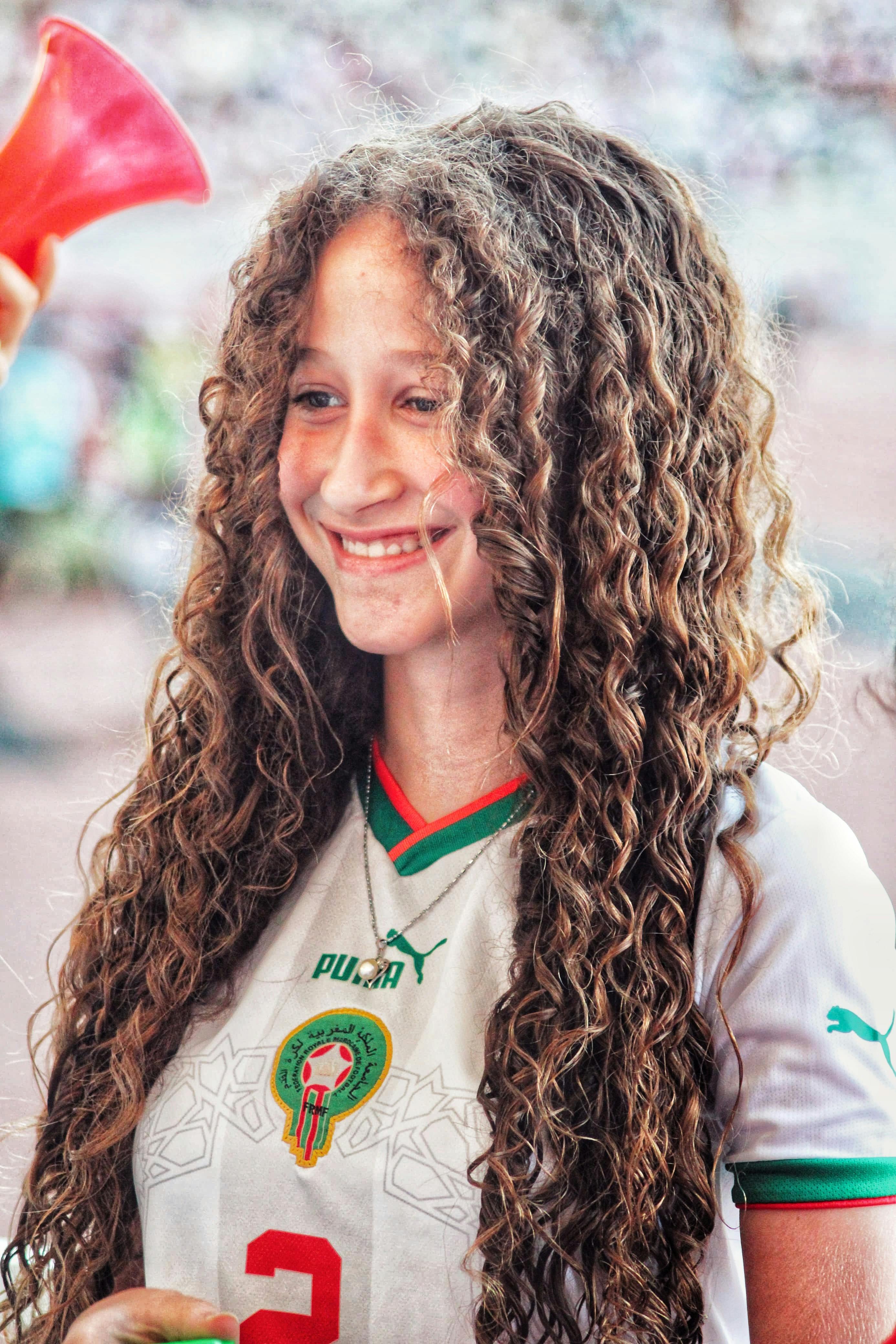 Teen girl in Moroccan soccer jersey smiling at outdoor event, showcasing fan spirit.