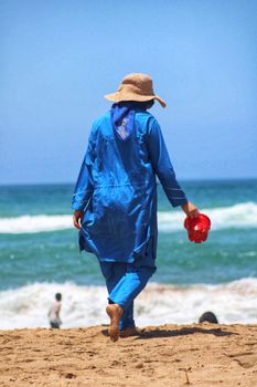 A woman in blue attire walks on a sandy beach holding red sandals during a sunny day.