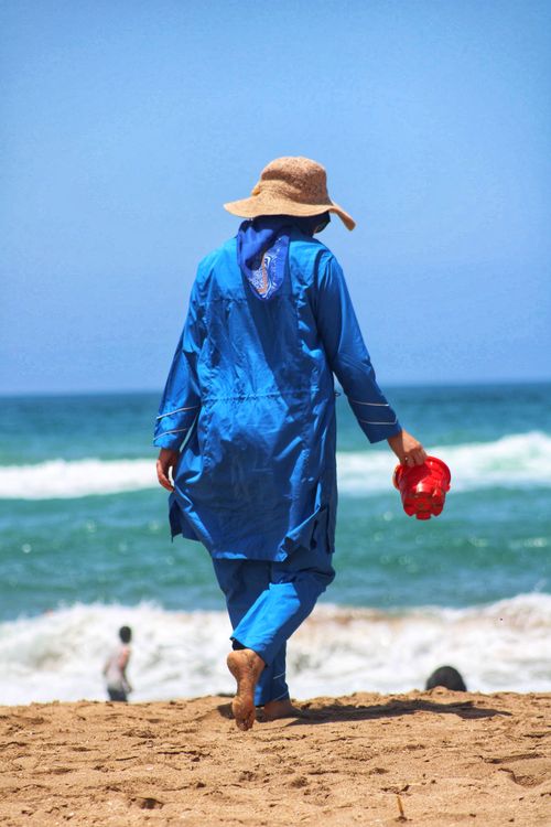 Free A woman in blue attire walks on a sandy beach holding red sandals during a sunny day. Stock Photo