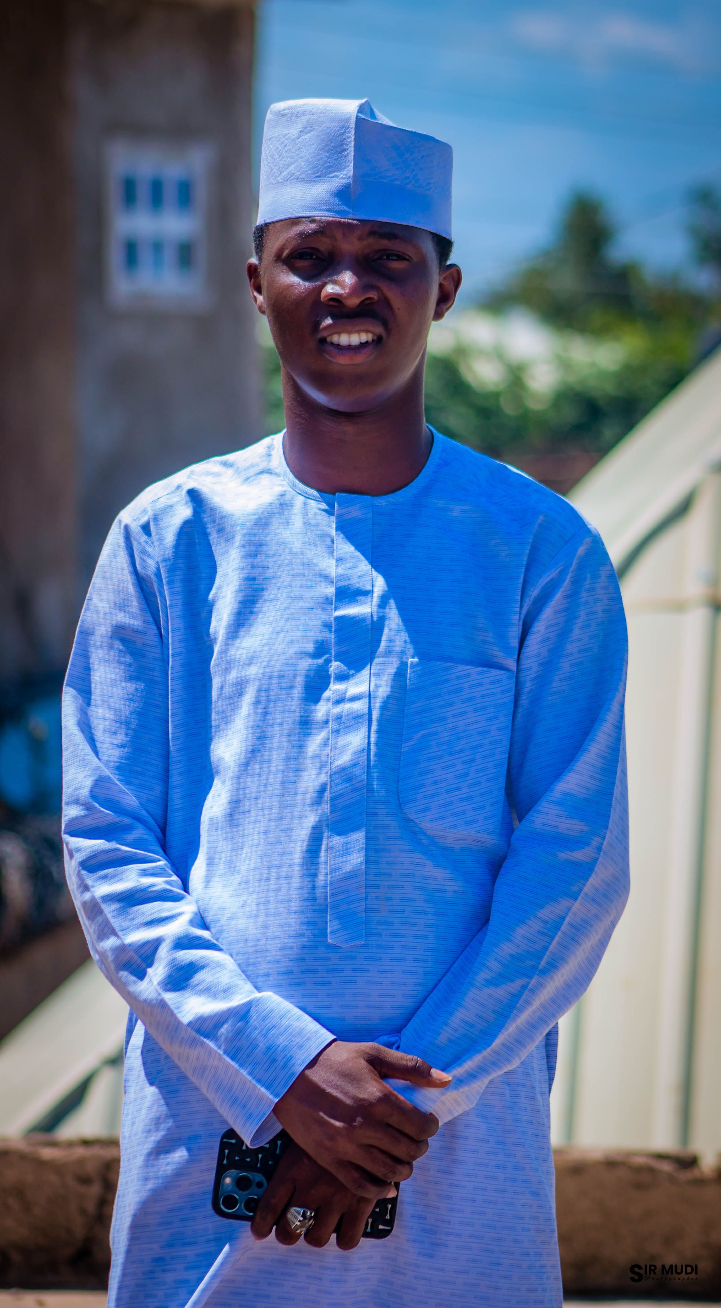 A young man in traditional attire confidently poses outdoors on a sunny day.
