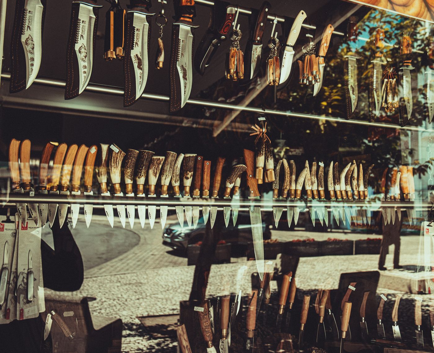 A variety of knives displayed in a shop window reflecting city street scene.