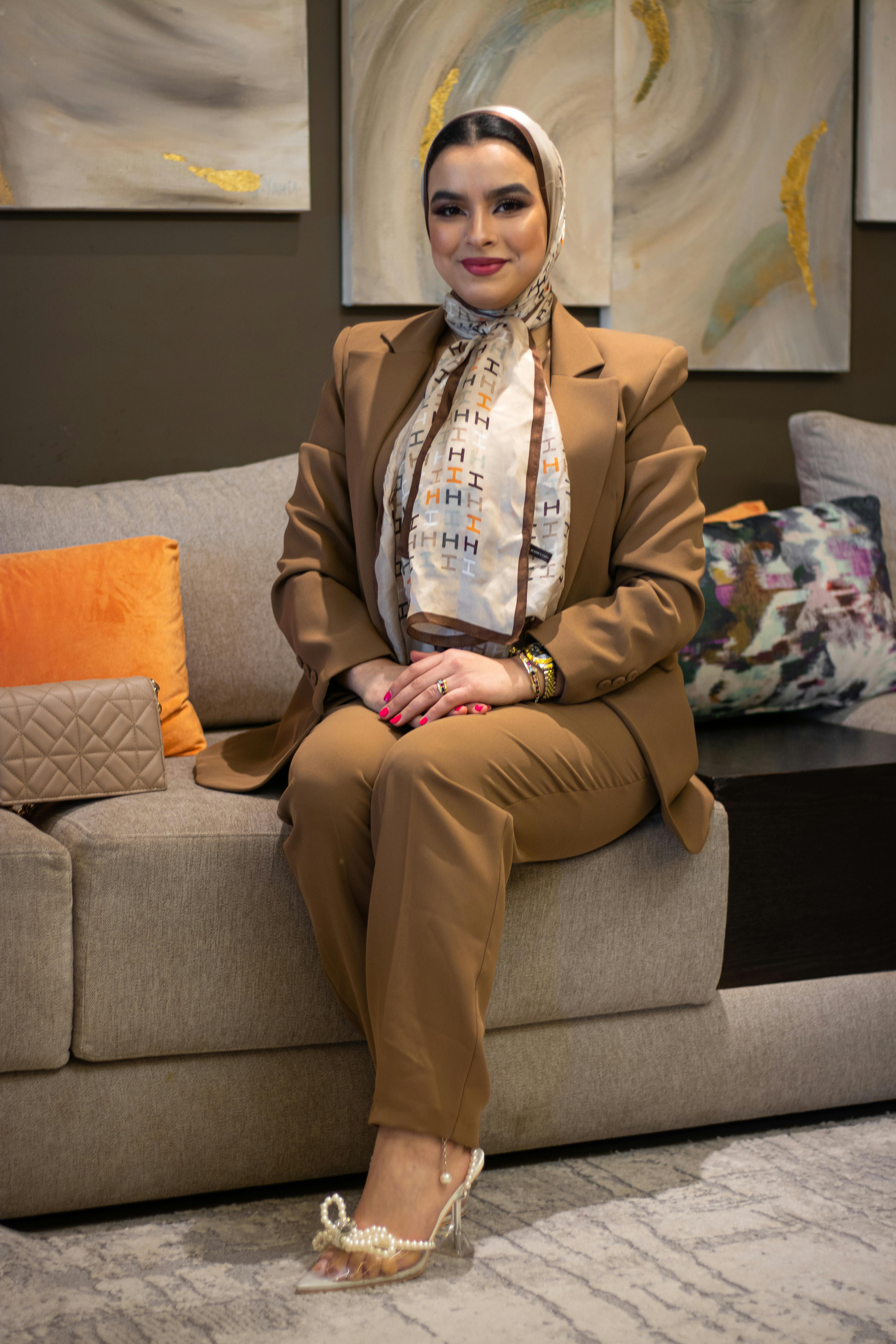 Stylish woman in a brown suit with a headscarf sitting on a modern sofa indoors.