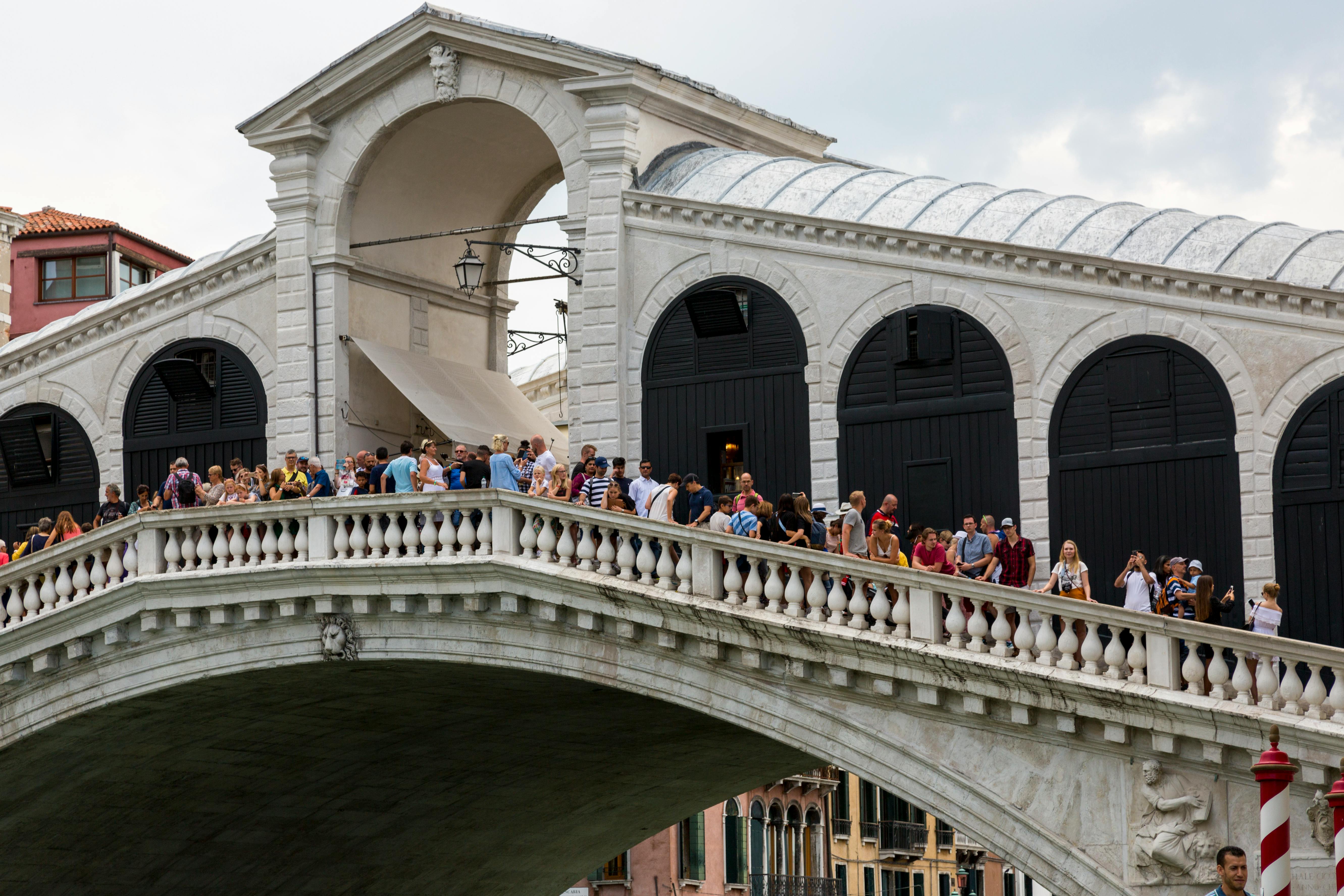 Crowd of people crossing a stone bridge beside a historic building with arched windows.
