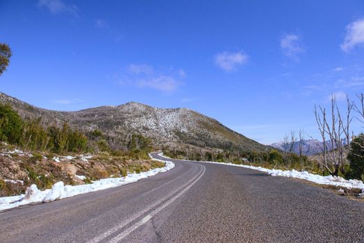 Winding road through snow-dusted mountains in Tasmania's wilderness under a clear blue sky.