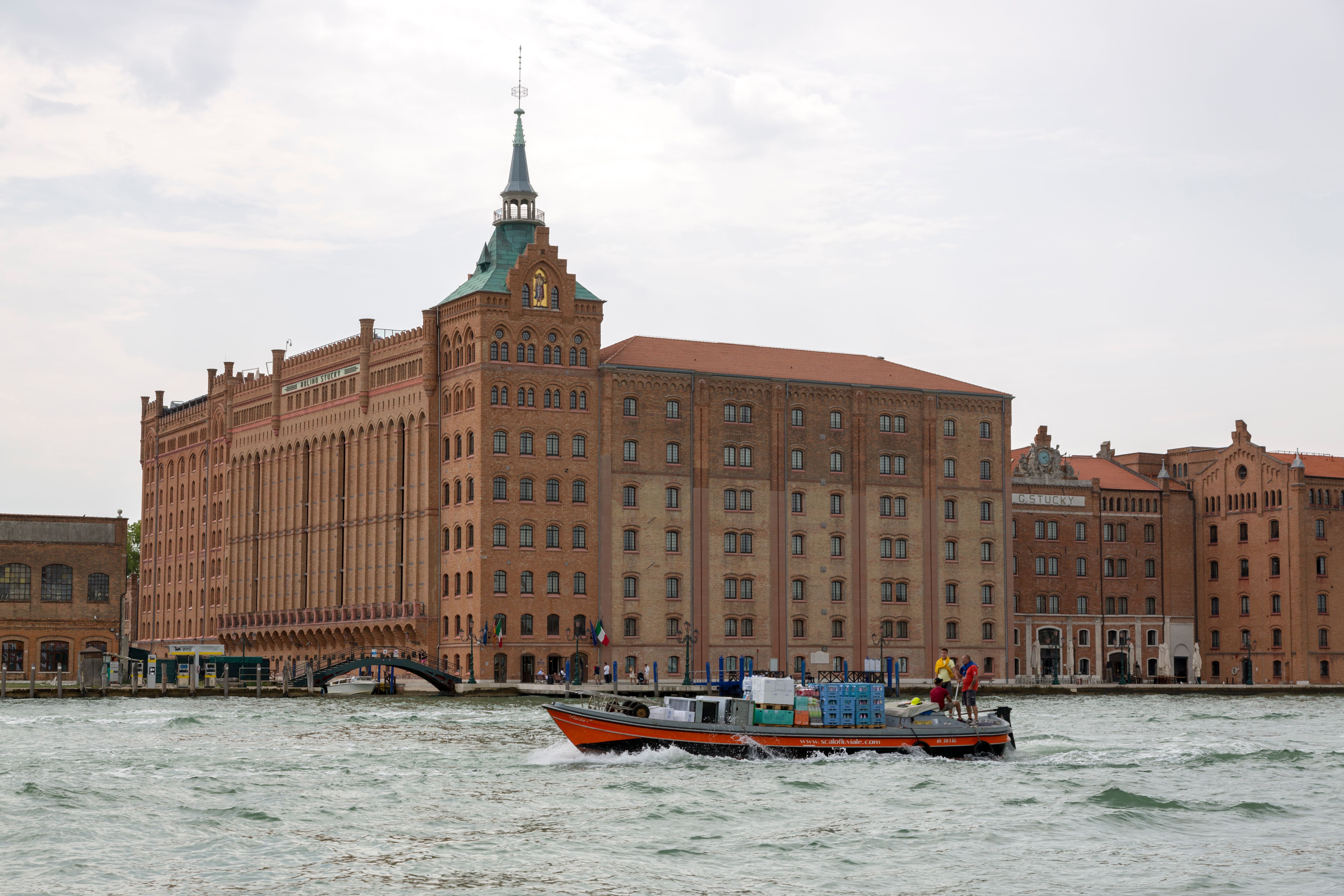 A very large brick building with a pointed green spire and a stepped roofline on the waterfront, with an orange work boat carrying crates on the water in front